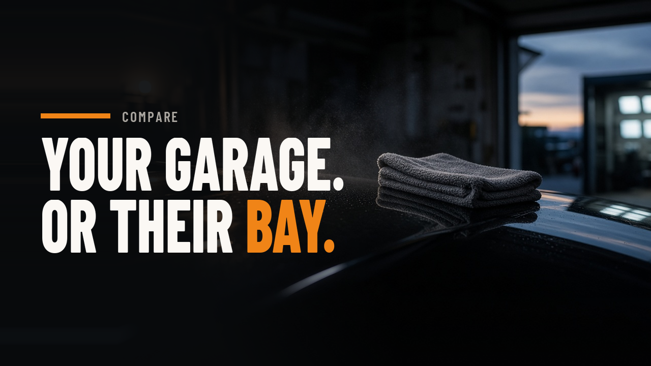 Clean dark car paint with a microfiber towel in a home garage at dusk, with a professional detailing bay lit up in the distance